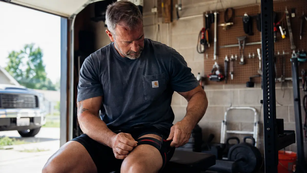 Gregory, a fifty-something mechanic, wrapping a BFR band around his upper leg in a garage gym with tools and a work bench in the background