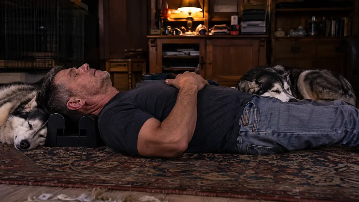Mechanic in his 50s lying on the floor of his living room using a cervical traction block under his neck, two huskies resting nearby