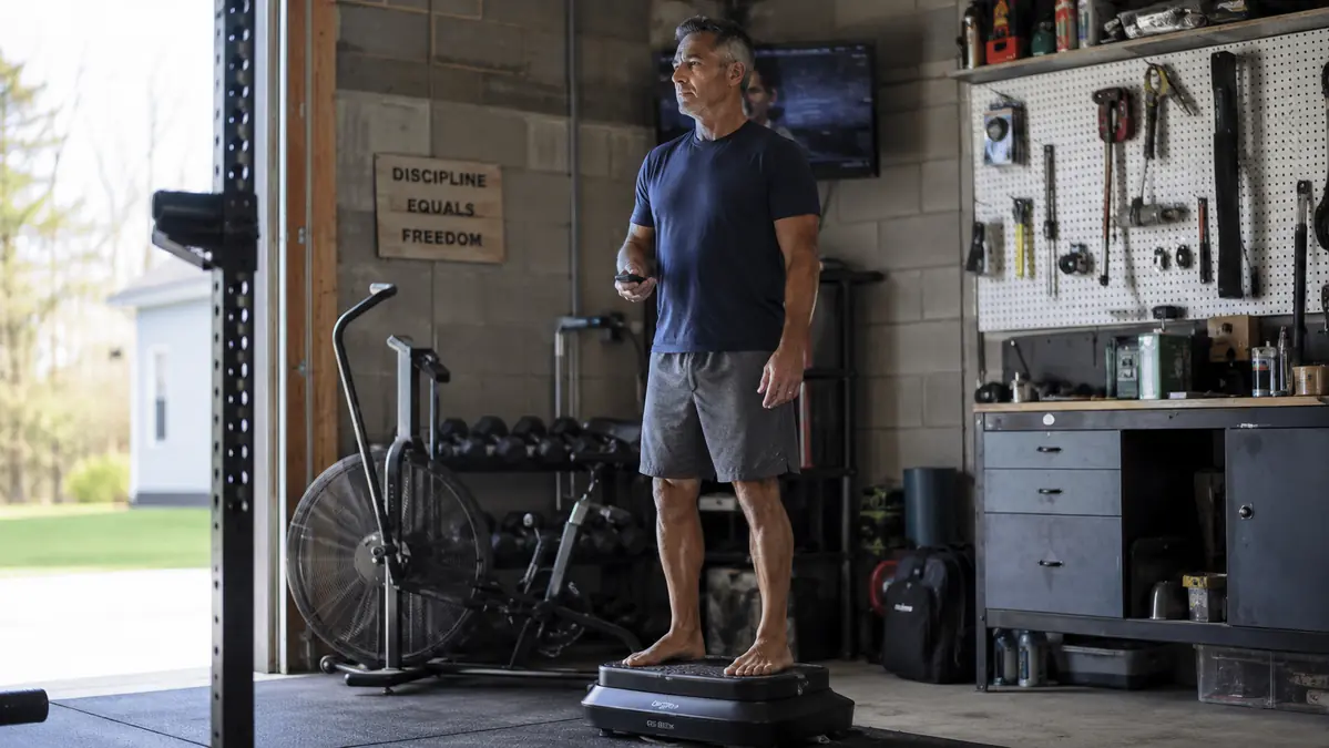 Gregory, a fifty-something mechanic, standing on a vibration plate in a garage gym with tools and a workbench in the background